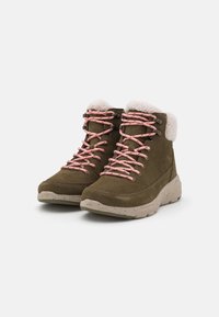 Une paire de bottes d'hiver en suède vert olive avec des lacets roses et une doublure en fausse fourrure blanche, semelles beige à taches, présentées sur un fond blanc.