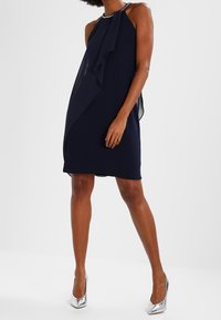 Robe halter bleu marine avec un vol d'eau cascade sur le devant, texture lisse et accents en métal argenté au niveau du décolleté. Associée à des talons argentés.