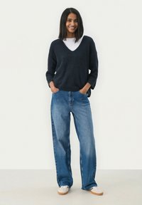 Pull en maille bleu marine avec un col en V, associé à un jean en denim léger bleu clair et ample. Chemise blanche en dessous et chaussures décontractées avec une semelle beige.