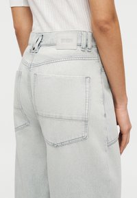 Lichtgrijze denim broek met een hoge taille, twee achterzakken en een zichtbaar merklintje. Gladde textuur met minimale stikseldetails.