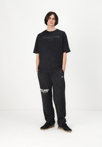 Melna oversize T-kreklu ar tekstu "Mūžīgi uz priekšu" un melnām teksturētām sporta biksēm ar "MCLAREN" logotipu, kombinēts ar melnām kedām.