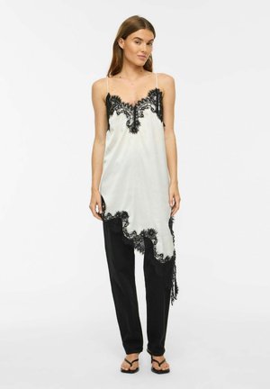Femme portant une robe asymétrique en soie blanche avec une bordure en dentelle noire par-dessus un pantalon noir et des talons ouverts noirs, debout sur fond blanc.