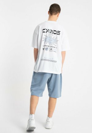 Mężczyzna stojący tyłem do widza, ubrany w białą koszulkę z nadrukiem „CHAOS” i niebieskim abstrakcyjnym wzorem, jasnoniebieskie spodenki, białe skarpetki oraz sportowe buty.