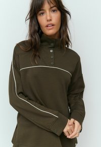 Sweatshirt vert foncé à col haut avec des accents et des boutons blancs sur le devant. Il présente une coupe décontractée et un tissu texturé.