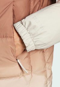Insulated jacket with a gradient color scheme from light beige to mauve, featuring a quilted texture and elastic cuffs. Includes a zippered pocket.