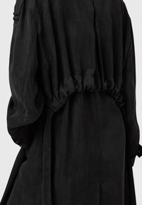 Svart plagg med avslappnad passform, markerad midja, vida ärmar och slät textur. Har ett knytbälte och en subtil glans.