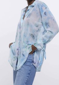 Sheer, light blue blouse with floral pattern, long sleeves tied at cuffs, and a button-down front. Features subtle horizontal texture.