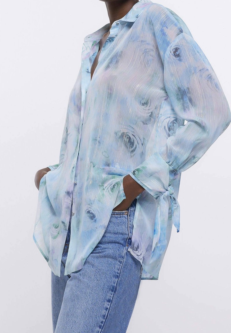 Sheer, light blue blouse with floral pattern, long sleeves tied at cuffs, and a button-down front. Features subtle horizontal texture.