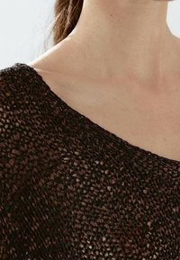 Gros plan sur l'épaule et le cou d'une personne portant un haut tricoté brun foncé, à mailles lâches, laissant entrevoir la peau en dessous.