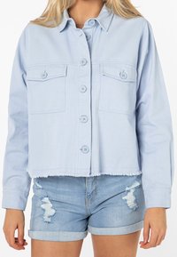 Světle modrá džínsová bunda s krátkým střihem, dvěma předními kapsami, zapínáním na knoflíky a roztrhaným lemem, doplněná o potrhané džínsové šortky.