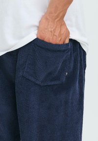 Main partiellement dans la poche arrière d'un pantalon en velours côtelé bleu marine porté avec une chemise blanche.