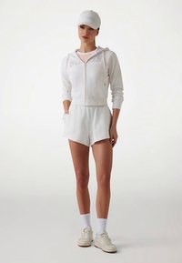 Sweat à capuche zippé en coton blanc avec logo, shorts assortis, casquette blanche et baskets. Les chaussettes sont blanches. Esthétique simple et sportive.
