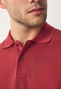 Polo rouge texturé avec un col à boutons, comprenant une petite poche poitrine et deux boutons à l'avant, mettant en valeur une structure en tissu côtelé.