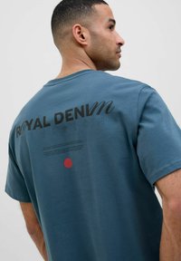 Zila kokvilnas T-kreklu ar apaļu kakla izgriezumu un īsām piedurknēm. Melno krāsā izcelta teksts "ROYAL DENIM DIVISION" un sarkanu punktiņu akcents.