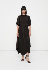 Lang, mørkebrun kjole med korte puffermer, samlet midje og asymmetrisk hem. Glatt tekstur, parret med svarte ankelrem sandaler.