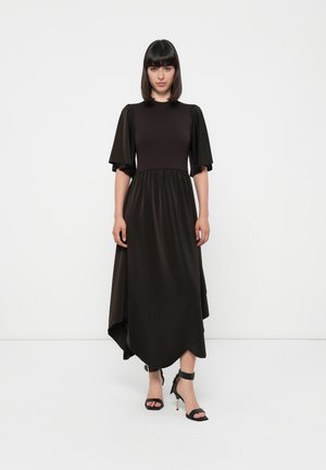 Long, dark brown dress with short puff sleeves, gathered waist, and asymmetrical hem. Smooth texture, paired with black ankle strap heels.