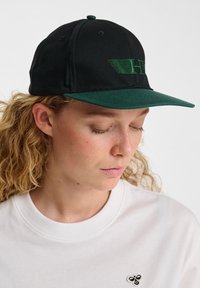 Black cap with a green visor featuring an embroidered logo. The fabric appears smooth with visible stitching details. Worn with a white sweatshirt.