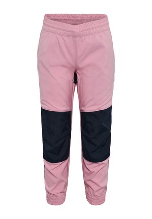 Lachend kind met lang blond haar dat een beige t-shirt draagt met outdoorprint, roze en zwarte broek en witte sneakers, staand met licht gespreide armen.
