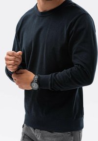 Felpa blu navy in cotone, con maniche lunghe, collo a giro e polsini a costine. Indossata con un orologio nero.