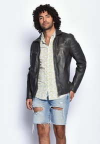 Schwarze Lederjacke mit gesteppten Ärmeln, kombiniert mit einem floral gemusterten Hemd und ausgewaschenen Denim-Shorts. Das Model steht selbstbewusst da.