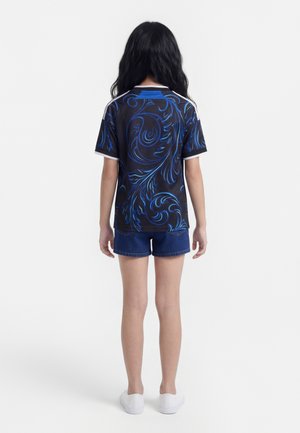 Meisje met lang zwart haar, een zwart shirt met blauwe draaipatronen, blauwe denimshorts en witte instapschoenen, staand met de rug naar de kijker.