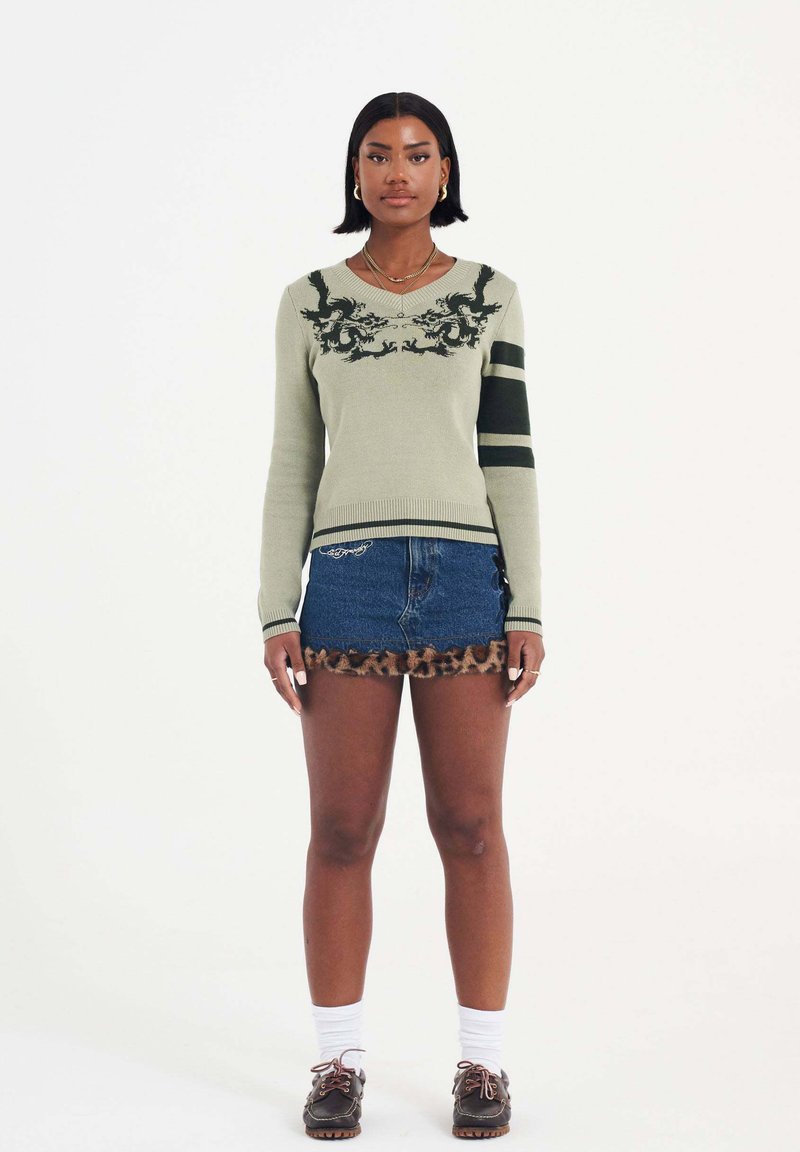 Grüner gestrickter Pullover mit schwarzem Drachendesign und gestreiften Ärmeln, kombiniert mit einem Jeansrock mit Leopardenmuster-Trim. Weiße Socken und braune Schuhe.