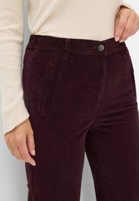 Bordeaux kordfløjspants med en struktureret overflade, der har frontlommer og en metalknaplukning i taljen.