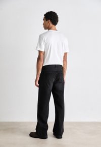 Schwarze Jeans im relaxed Fit, kombiniert mit einem schlichten weißen T-Shirt aus Baumwolle. Die Figur wird von hinten gezeigt, die Hände an den Seiten.
