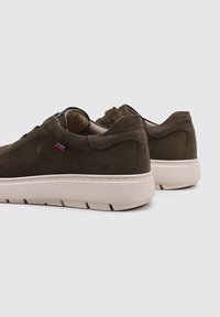 Zapatillas de ante verde oliva con una suela de goma crema, que presentan líneas simples y un diseño minimalista. Con un pequeño logo en una etiqueta.