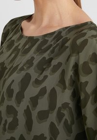 Top verde con escote redondo que presenta un patrón de camuflaje oscuro. La tela parece suave con tonos de verde ligeramente variados.