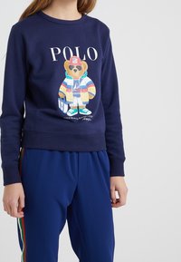 Sweat-shirt bleu marine avec un graphique d'ours vêtu d'une tenue colorée. Présente des poignet et un ourlet côtelés, associé à un pantalon de jogging bleu avec des rayures multicolores sur les côtés.