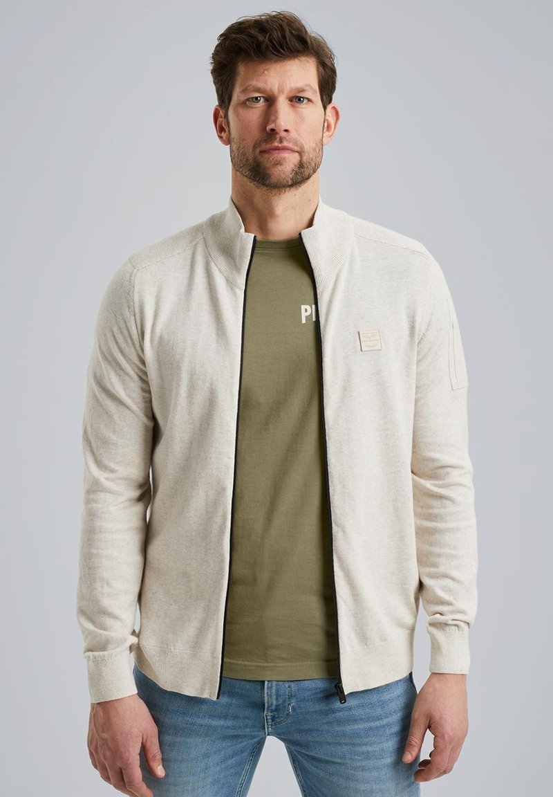 Hellbeige gesmokte Strickjacke aus weichem Material, mit hohem Kragen, kontrastierendem schwarzen Reißverschluss und kleinem Logo-Patch auf der linken Brust.