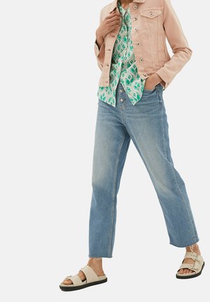 Hellrosa Jeansjacke, gemustertes grünes Hemd, hellblaue gerade Jeans und beigefarbene Sandalen mit schwarzen Sohlen und Akzenten.