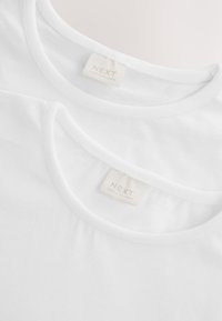 Zwei weiße Baumwoll-T-Shirts übereinandergelegt, mit rundem Ausschnitt und sichtbaren Etiketten mit der Aufschrift "NEXT", hergestellt in Sri Lanka. Glatte Textur.