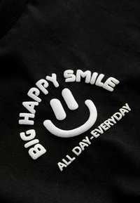 Black fabric with white embroidered text that reads "BIG HAPPY SMILE" above a smiling face and "ALL DAY EVERYDAY" below. Soft texture.