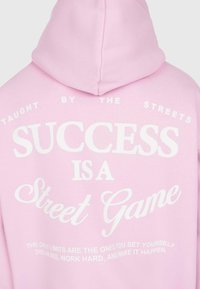 Felpa rosa con testo bianco che recita "Insegnato dalle strade, il successo è un gioco di strada" e una citazione motivazionale in fondo.