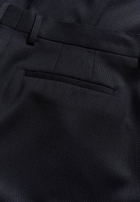 Tmavě modré kalhoty s texturovanou tkaninou. Mají vypasovaný pas a jednu zadní kapsu s úzkým otvorem.