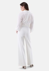 Blusa bianca con maniche fluttuanti e colletto alto, abbinata a pantaloni bianchi a gamba larga e scarpe con tacco blocco di colore chiaro.