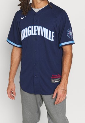 Homme portant un maillot de sport Wrigleyville bleu marine avec des bordures bleu clair et un pantalon de survêtement gris, debout devant un fond uni.