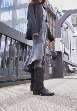 Una persona con stivali neri fino a metà polpaccio, leggings grigi, una gonna di denim sfilacciata e una giacca nera si trova su un marciapiede urbano vicino a una ringhiera metallica.