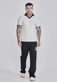 Bela polo majica s kratkimi rokavi z črnimi črtami in črnim ovratnikom, v kombinaciji s črnimi hlačami z belim logotipom. Model stoji, tetovaže so vidne.