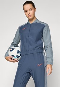 Nike sportssæt i navy og grå med en zip-jakke, elastisk talje og logo-detaljer. Holder en mønstret fodbold.