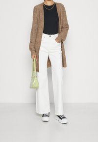 Brązowy sweter na guziki noszony na czarnej koszuli, zestawiony z białymi spodniami o szerokich nogawkach oraz czarnymi trampkami. Stylizację uzupełnia zielona torebka o teksturowanej powierzchni.