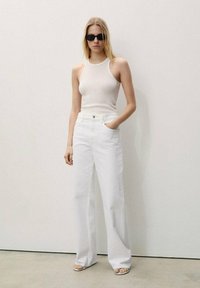 Hvide højtaljede jeans med bred ben design, parret med en tætsiddende, ribbet cremefarvet tanktop. Model iført sorte solbriller og minimalistisk fodtøj.