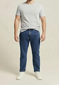 Homme portant un t-shirt gris clair à manches courtes, un jean bleu avec les revers retroussés et des baskets blanches, debout devant un fond neutre.
