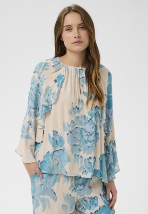 Mujer con blusa beige y azul de flores con volantes y pantalones a juego, de pie con una mano en el bolsillo sobre fondo blanco.