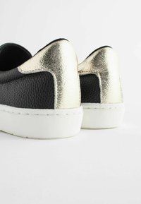 Chaussures en cuir noir à enfiler avec une surface texturée, présentant des accents métalliques dorés au talon et une semelle en caoutchouc blanche.