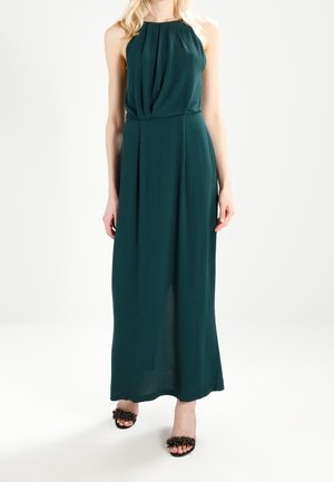 Femme portant une robe maxi sans manches vert foncé à col halter avec des plis, associée à des sandales noires à bout ouvert avec des clous décoratifs.