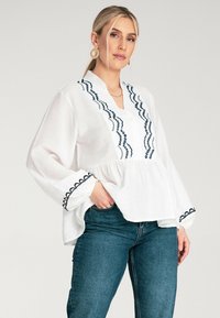 Biała bluzka z niebieskimi haftowanymi detalami wzdłuż dekoltu i mankietów, o luźnym kroju i długich rękawach, w połączeniu z ciemnoniebieskimi dżinsami.