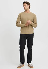 Lichtbeige gebreide trui met lange mouwen, ronde halslijn en geribde zoom, gedragen met zwarte jeans en grijzige sportschoenen.